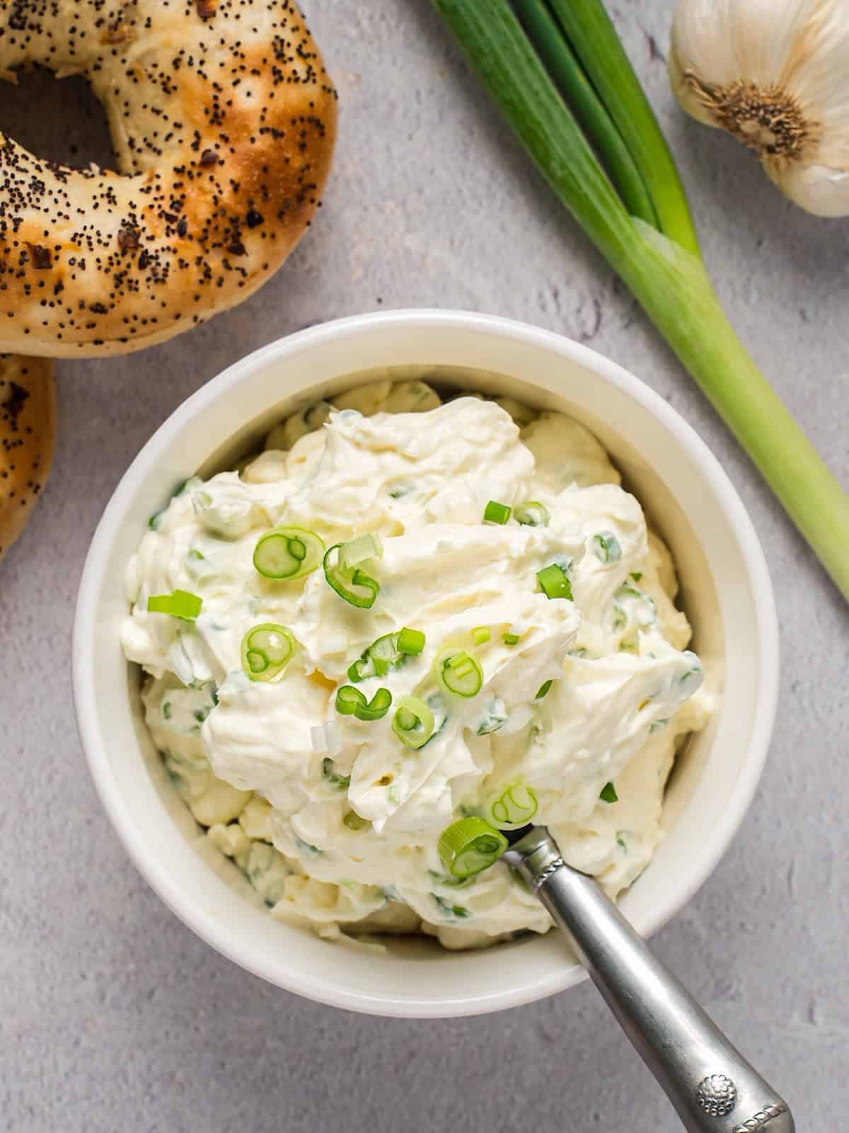 Scallion cream cheese spread in a bowl plated near everything bagel, scallion, and garlic bulb.