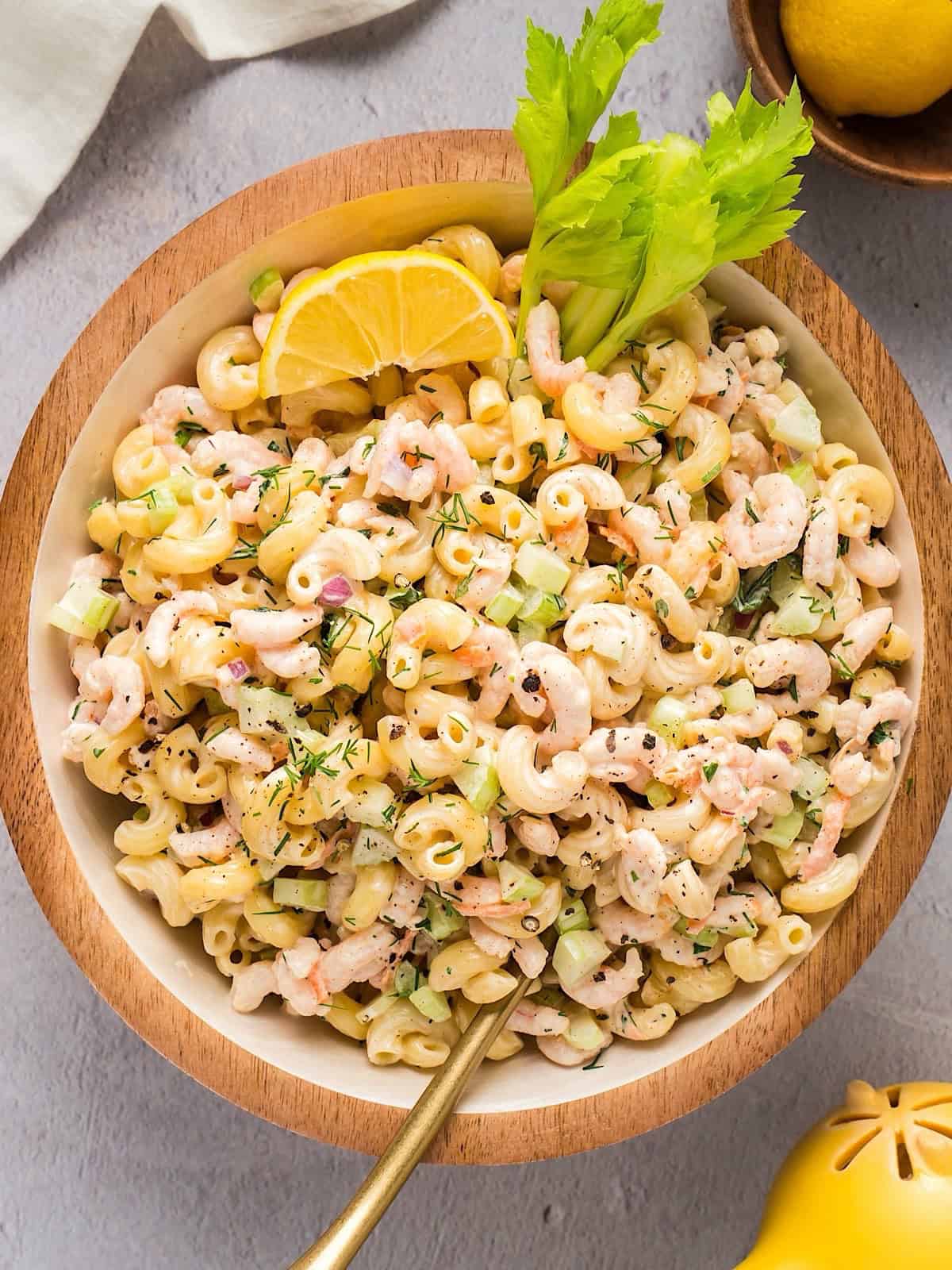 Creamy shrimp macaroni salad in a wooden bowl with lemon wedge, celery stick, and composition elements (lemon squeezer, linen, etc)