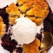 A gold spoon dipping into Southern blackberry cobbler topped with vanilla ice cream.