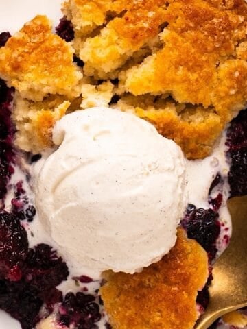 A gold spoon dipping into Southern blackberry cobbler topped with vanilla ice cream.
