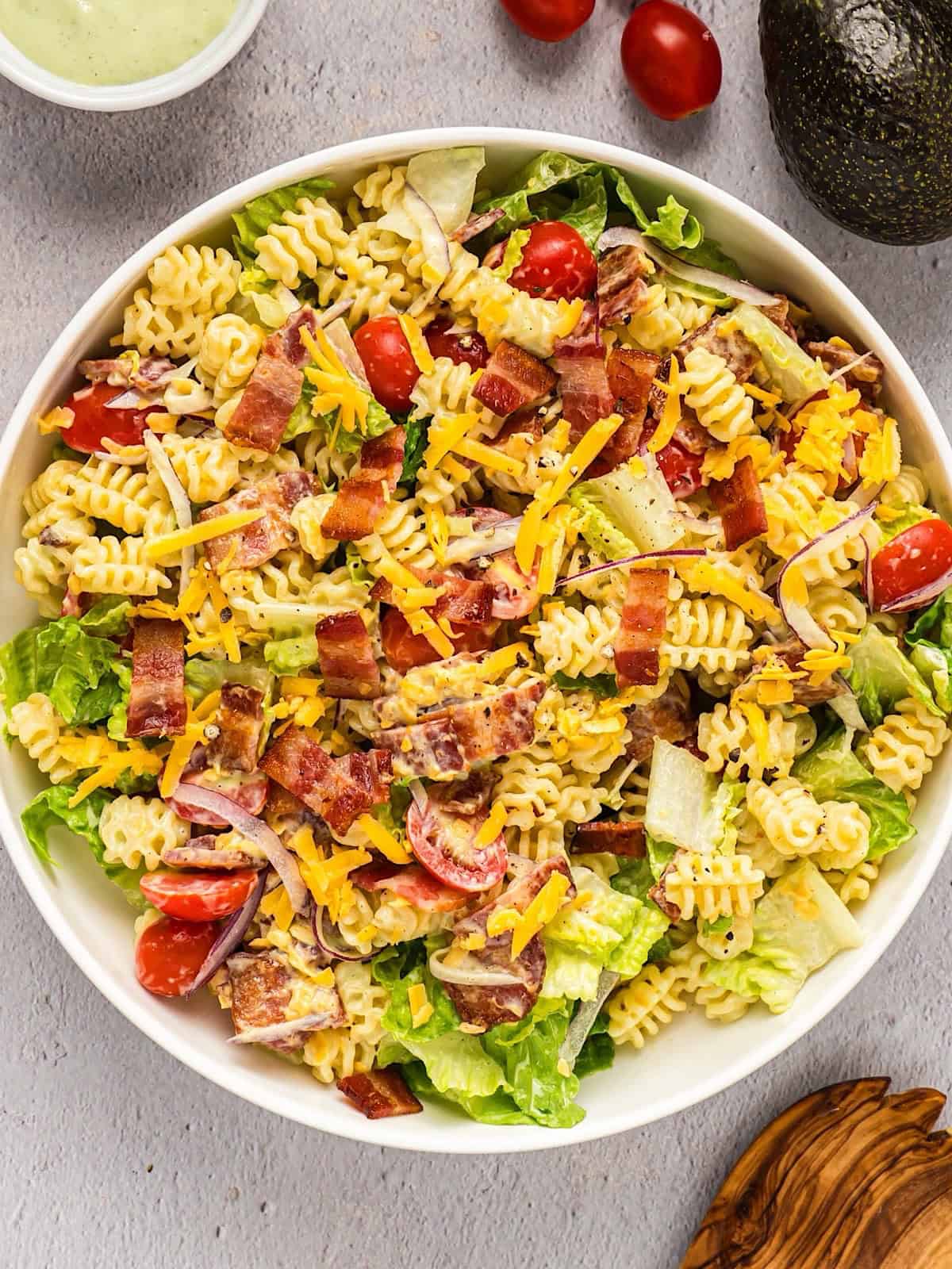 BLT pasta salad tossed in a wide serving bowl and plated near avocado, tomato, and wooden spoon.
