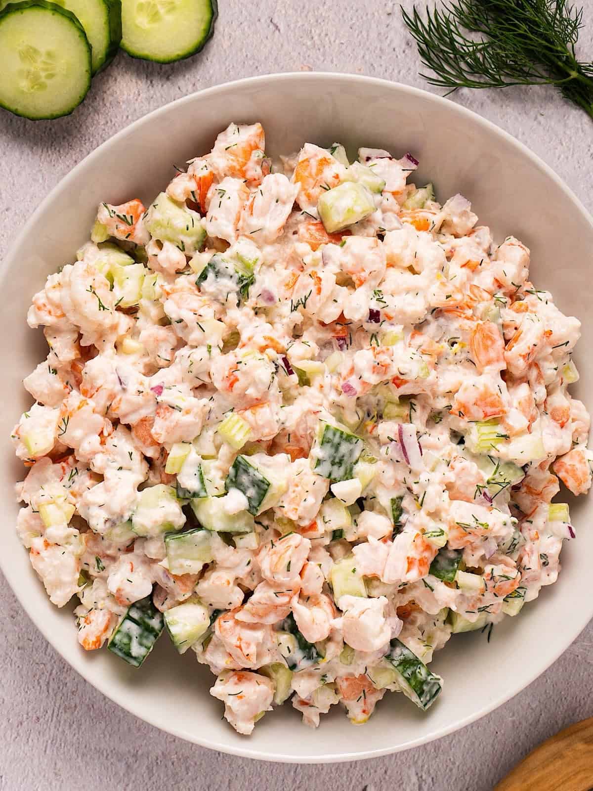Creamy cucumber shrimp salad with Greek yogurt in a white bowl plated near cucumbers, fresh dill, and a wooden spoon.