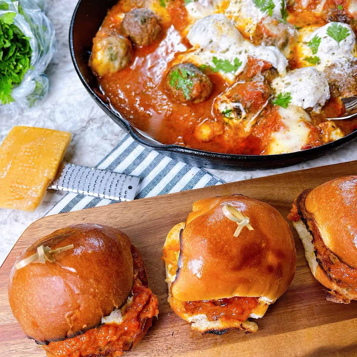 Prepared meatball sliders on a serving board with skillet in background.