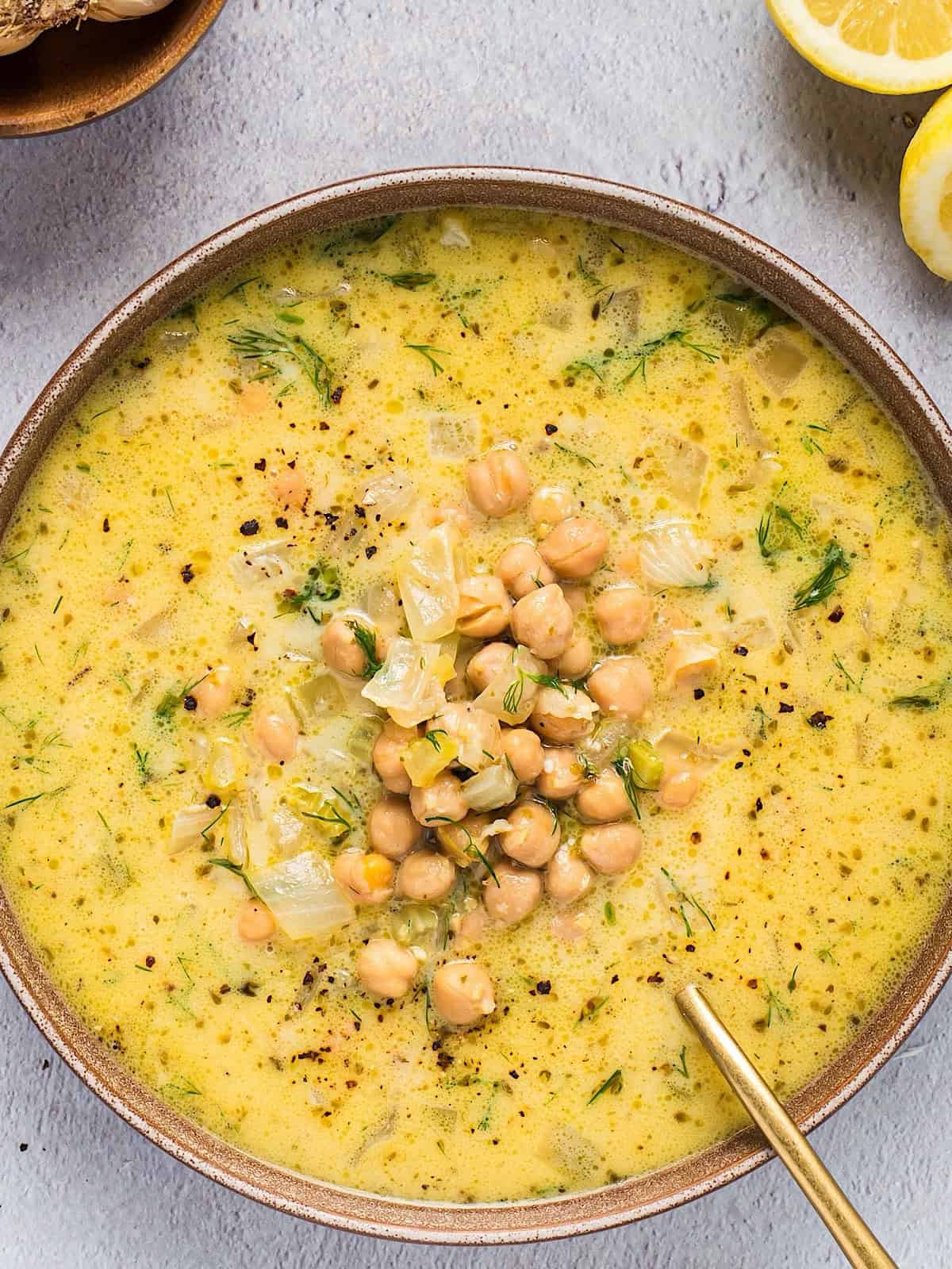 A wide bowl of Greek chicken soup with lemon plated near lemons and bowl of garlic.