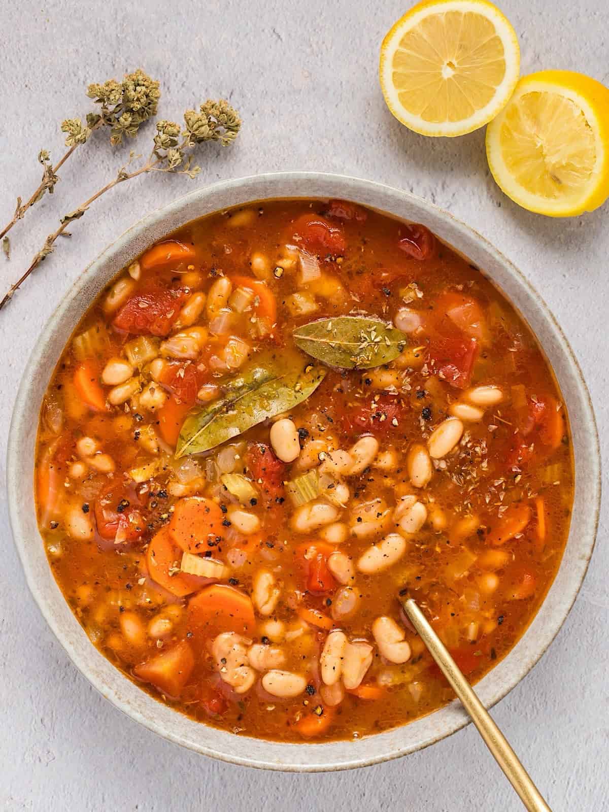 A wide bowl of Greek fasolada soup plated near lemon and dried oregano stems.