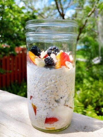 A mason jar of peaches and cream overnight oats with toppings sitting on a deck with sunshine highlighting ingredients.
