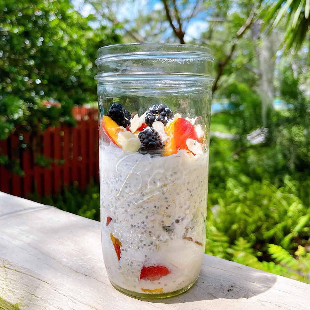 A mason jar of peaches and cream overnight oats with toppings sitting on a deck with sunshine highlighting ingredients.