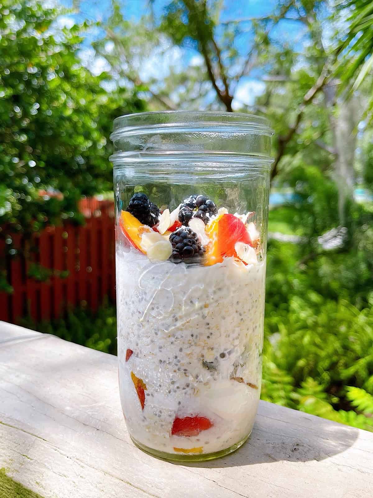 A mason jar of peaches and cream overnight oats with toppings sitting on a deck with sunshine highlighting ingredients.