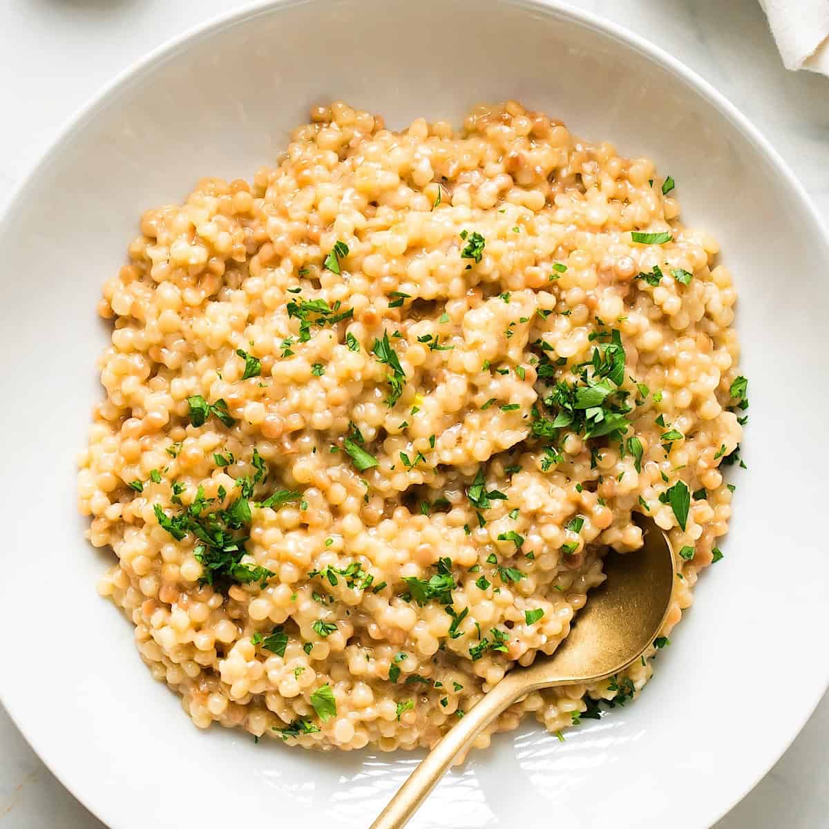 Garlic parmesan couscous with parsley garnish in a white dish with gold spoon.