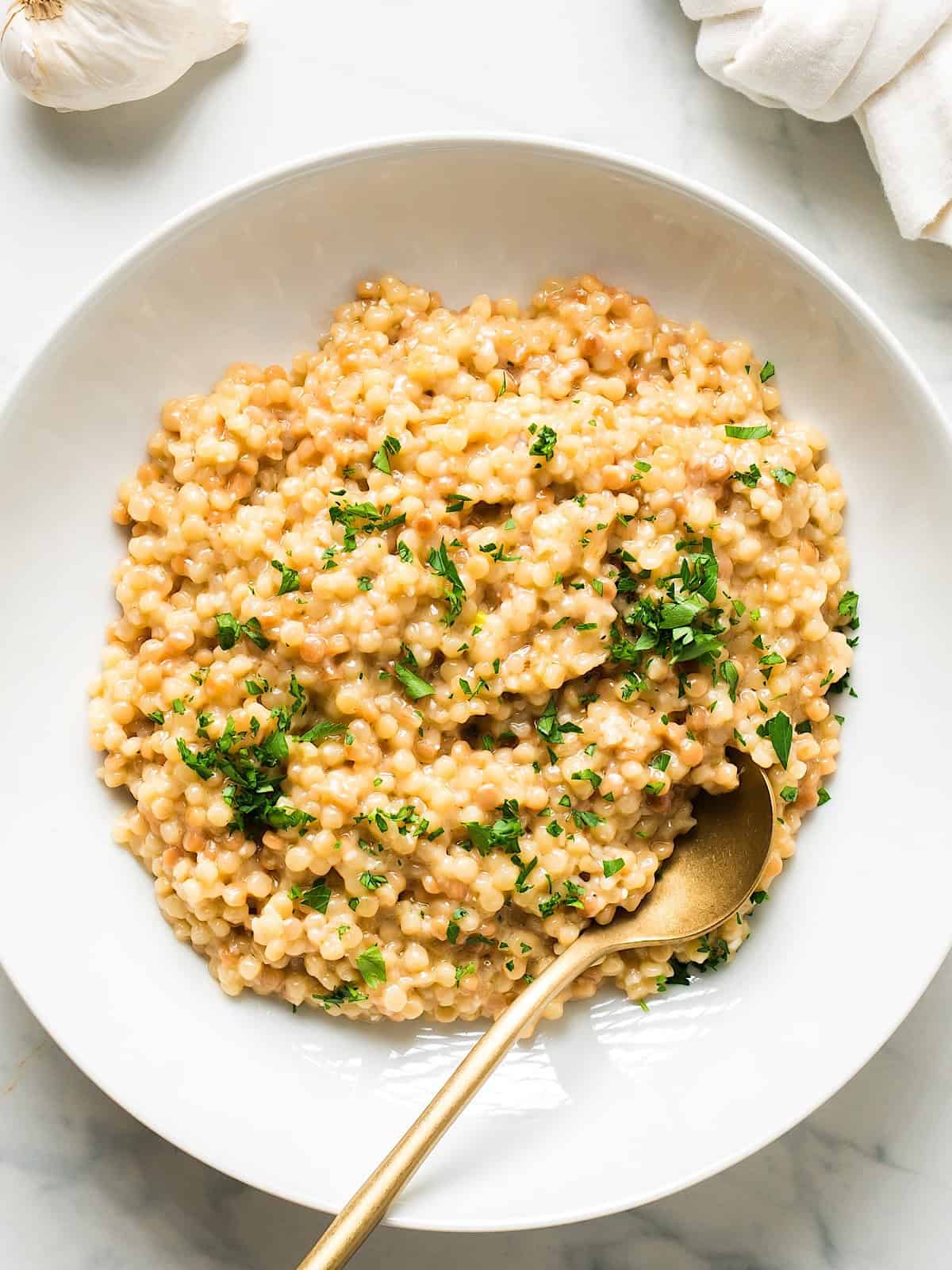 Garlic parmesan couscous with parsley garnish in a white dish with gold spoon.
