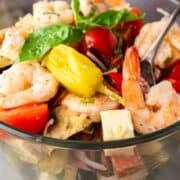 Tossed shrimp antipasto salad layered in glass serving dish with fresh basil leaves on top.