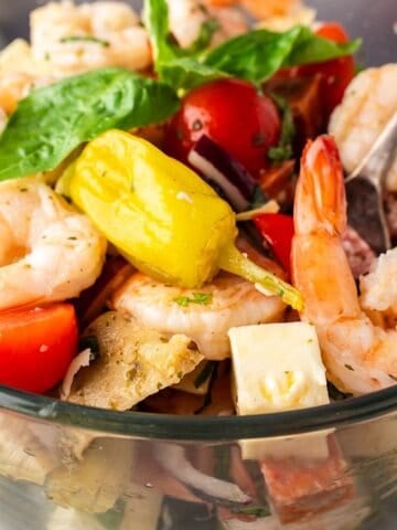 Tossed shrimp antipasto salad layered in glass serving dish with fresh basil leaves on top.