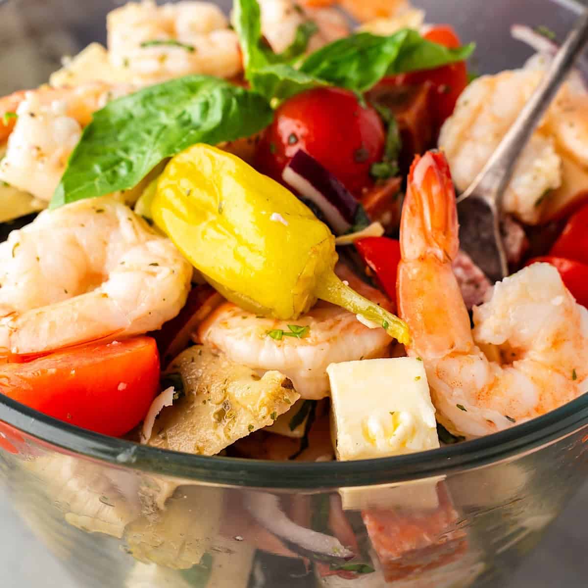 Tossed shrimp antipasto salad layered in glass serving dish with fresh basil leaves on top.