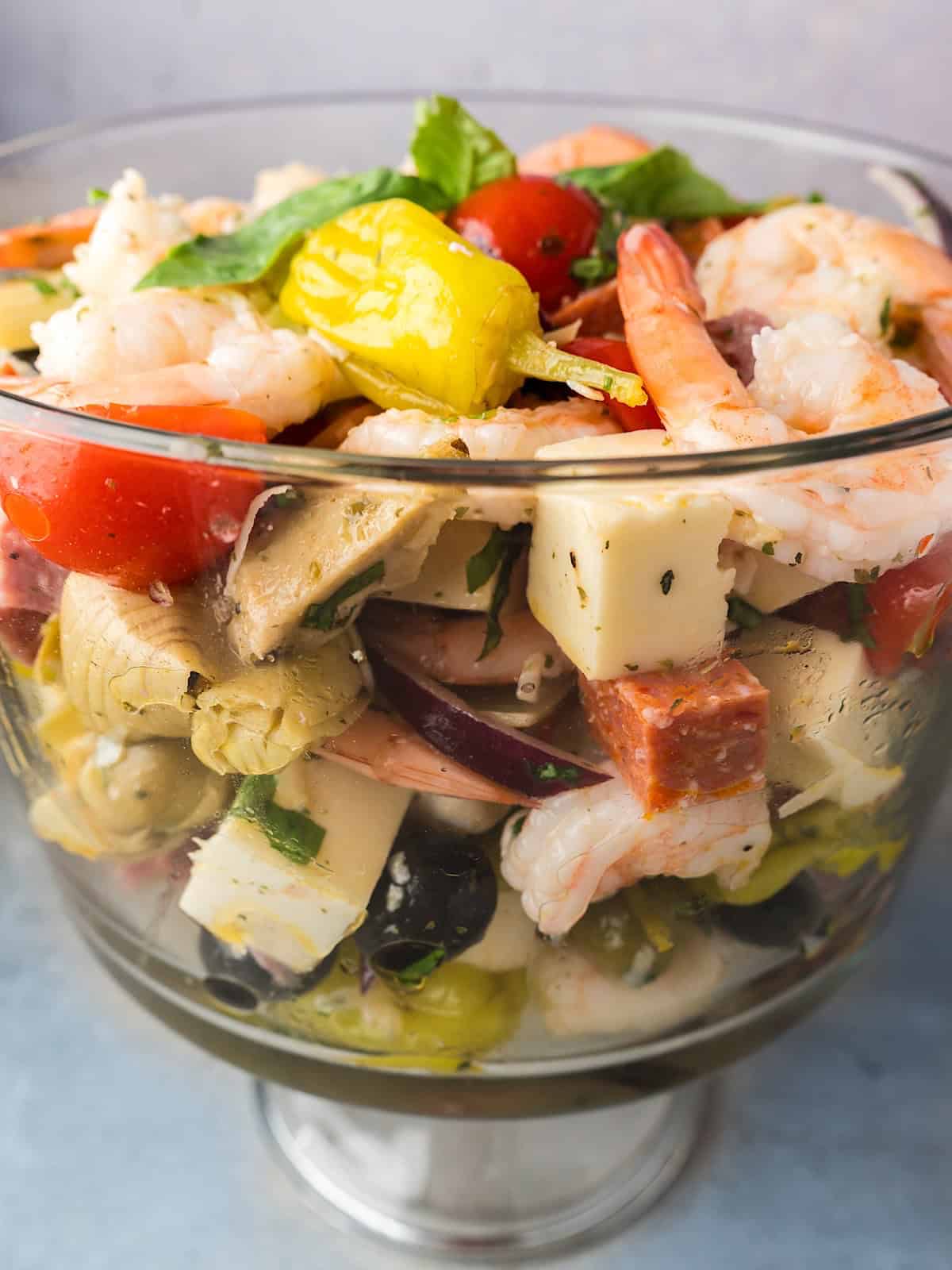 Tossed shrimp antipasto salad layered in glass serving dish with fresh basil leaves on top.