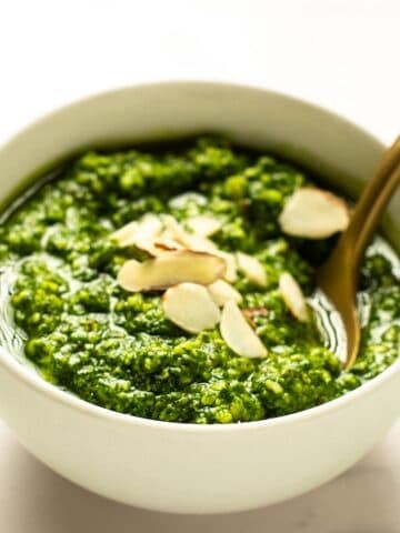 Basil pesto without pine nuts in a small white bowl with sliced almonds on top and a gold spoon.