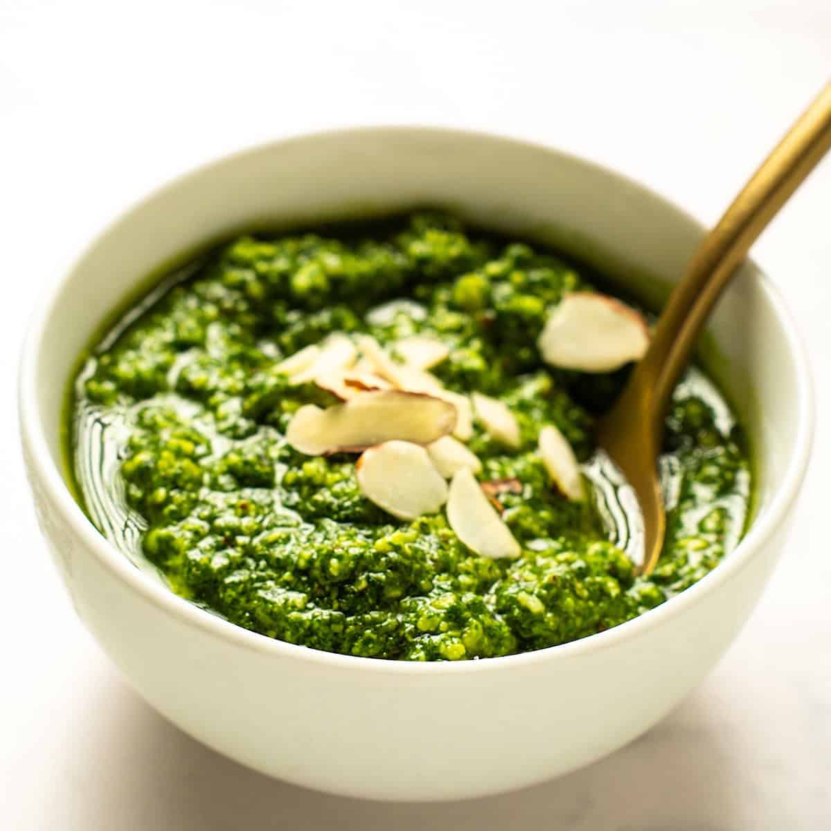 Basil pesto without pine nuts in a small white bowl with sliced almonds on top and a gold spoon.