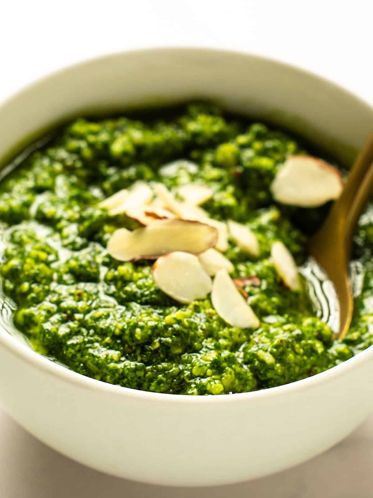 Basil pesto without pine nuts in a small white bowl with sliced almonds on top and a gold spoon.