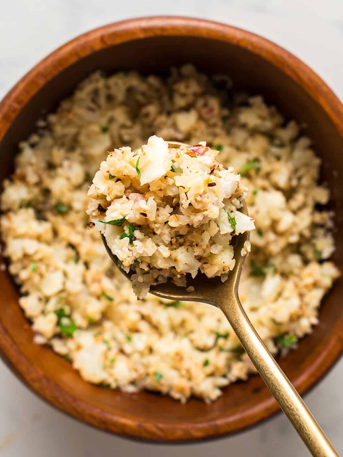 A gold spoon with lemon herb cauliflower rice being held over wooden bowl.