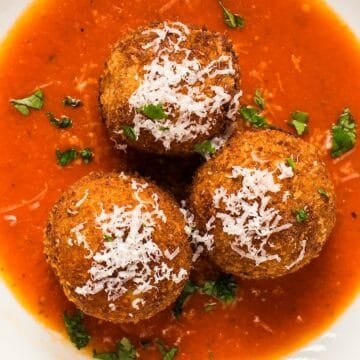 Three garnished arancini plated in a wide shallow bowl with marinara sauce.