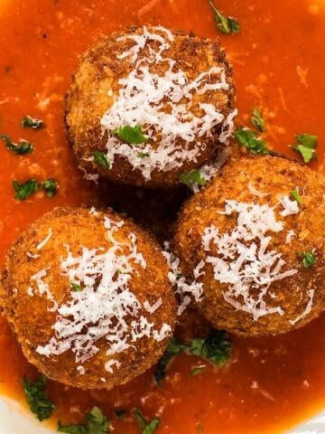 Three garnished arancini plated in a wide shallow bowl with marinara sauce.