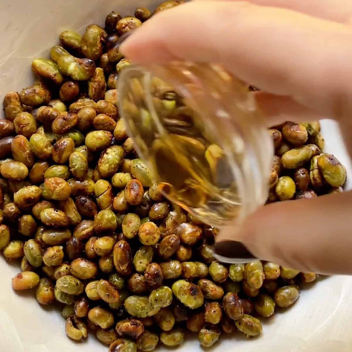 Drizzling sesame oil over roasted edamame in a wooden serving bowl.