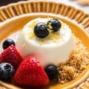 Lemon panna cotta on decorative plate garnished with berries, zest, and graham cracker crumbs.