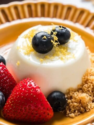 Lemon panna cotta on decorative plate garnished with berries, zest, and graham cracker crumbs.
