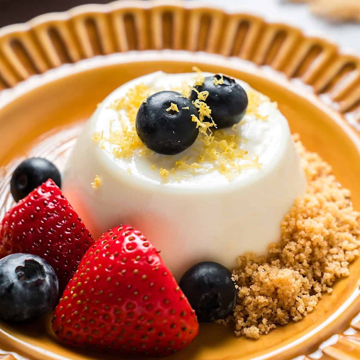 Lemon panna cotta on decorative plate garnished with berries, zest, and graham cracker crumbs.