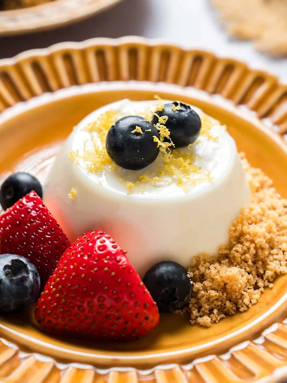 Lemon panna cotta on decorative plate garnished with berries, zest, and graham cracker crumbs.