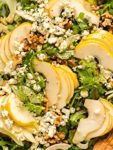 Pear arugula salad with gorgonzola, fennel, and walnuts on elegant platter with wooden serving utensil.