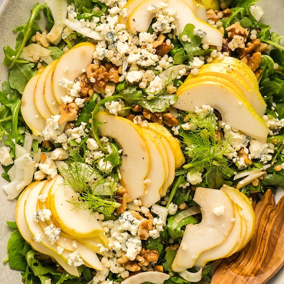 Pear arugula salad with gorgonzola, fennel, and walnuts on elegant platter with wooden serving utensil.