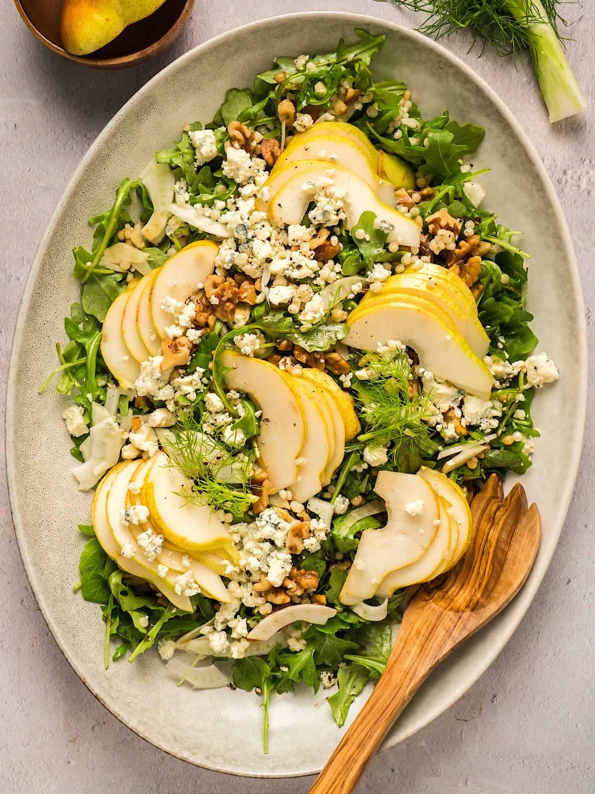 Pear arugula salad with gorgonzola, fennel, and walnuts on elegant platter with wooden serving utensil.