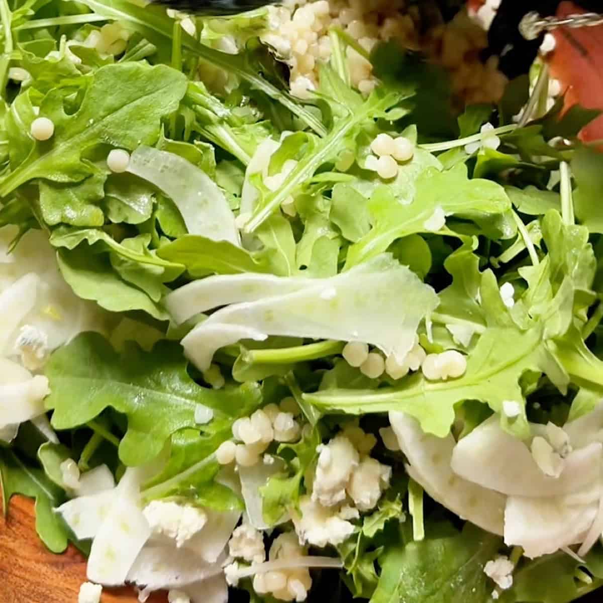 Tossing the arugula leaves with pearl couscous, fennel, blue cheese, and dressing.