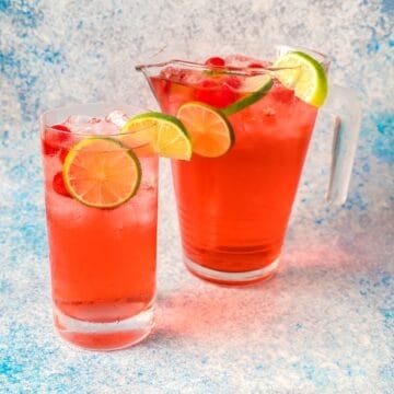 A glass of shirley temple with lime wheel, maraschino cherries, and cocktail ice next to a filled pitcher.