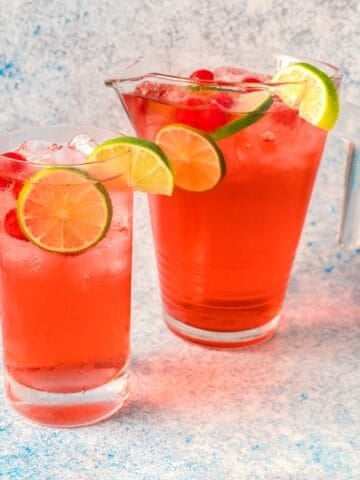 A glass of shirley temple with lime wheel, maraschino cherries, and cocktail ice next to a filled pitcher.