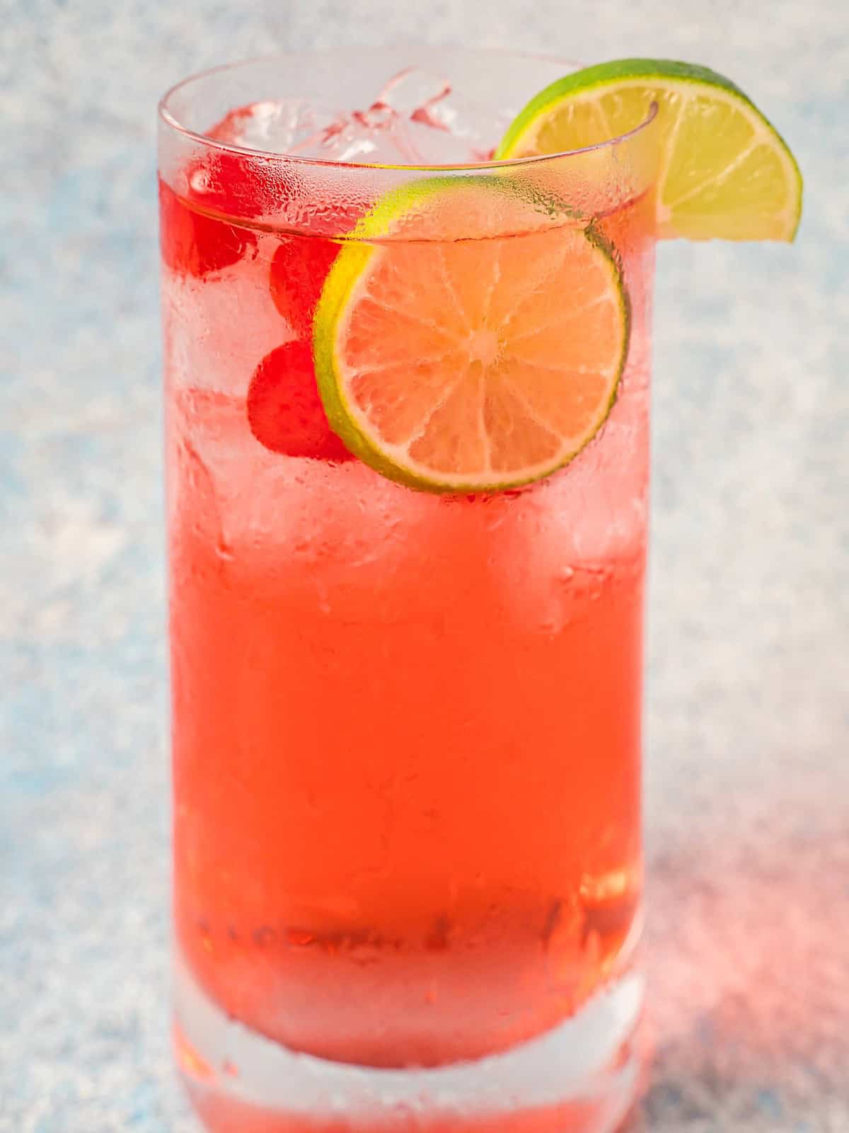 A tall glass of shirley temple with lime wheel, maraschino cherries, and cocktail ice.